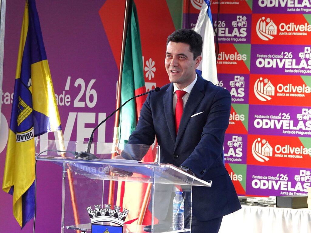 Homem de terno azul e gravata vermelha a proferir discurso numa tribuna, com fundo roxo e várias bandeiras.