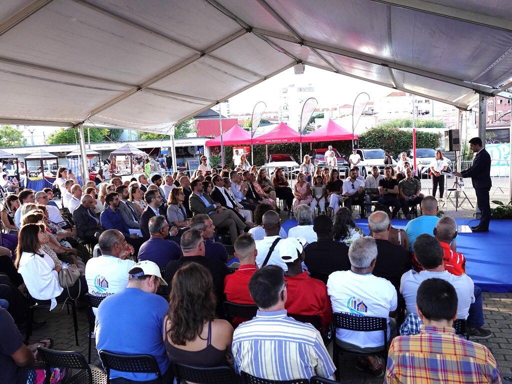 Palestra ao ar livre sob tenda, grupo de pessoas sentadas em círculo assistindo apresentador.