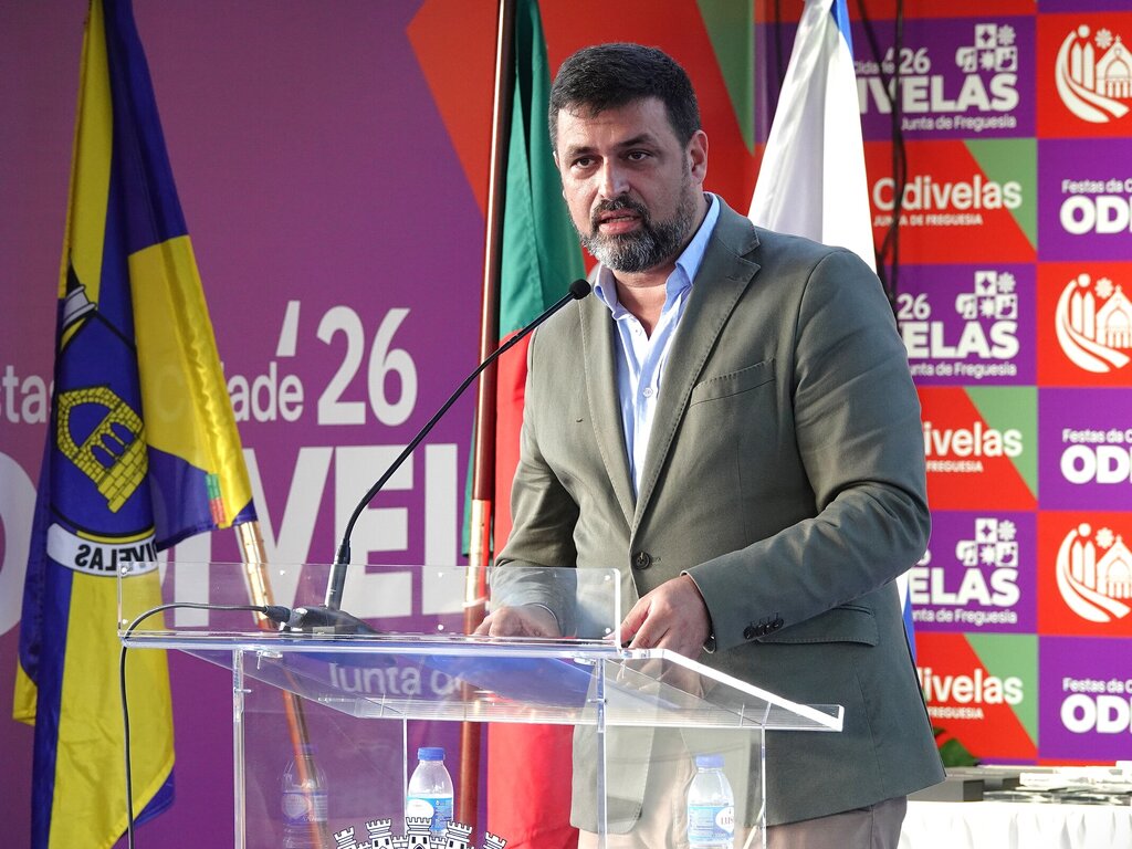 Homem de traje cinzento fala em púlpito transparente numa conferência, rodeado por bandeiras e um fundo colorido com logos.