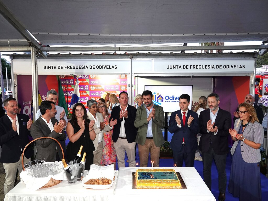 Grupo de pessoas a aplaudir ao lado de um bolo decorado, em evento institucional.